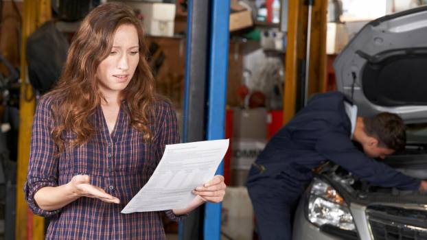 Woman holds Lexus dealership repair estimate in shock while mechanic works in the background.