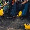 Row of factory workers with their helmets on the ground while they sit against a wall.