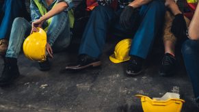 Row of factory workers with their helmets on the ground while they sit against a wall.