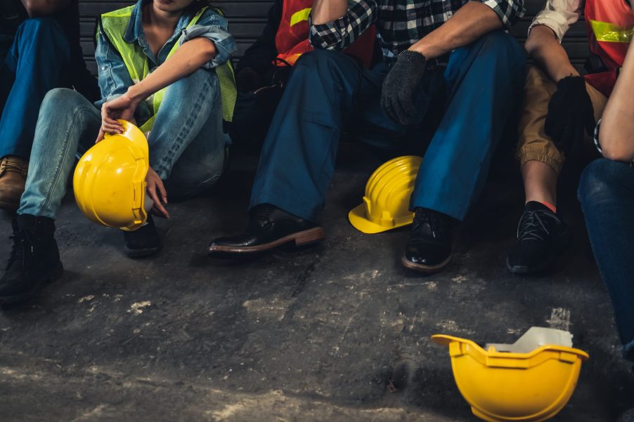 Row of factory workers with their helmets on the ground while they sit against a wall.