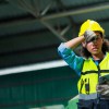 Factory worker wipes sweat off her forehead, a steel building visible in the background.