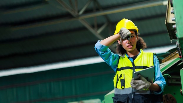 Factory worker wipes sweat off her forehead, a steel building visible in the background.