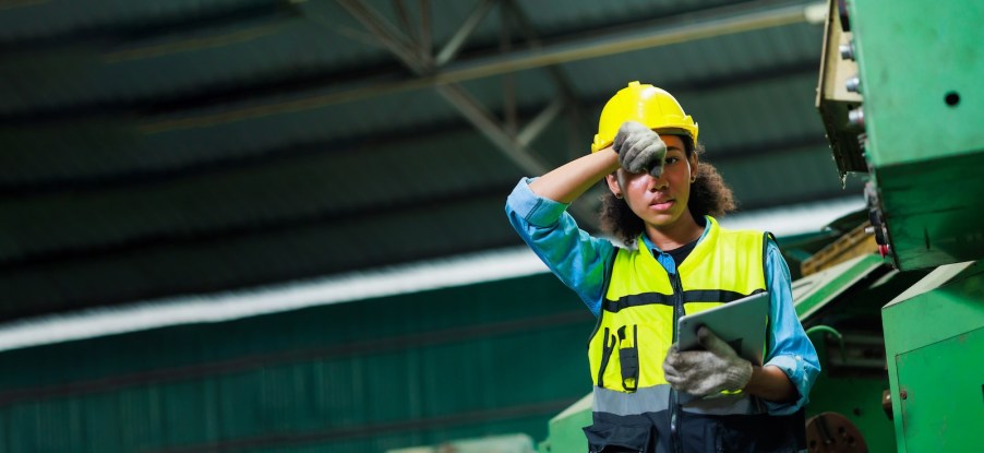 Factory worker wipes sweat off her forehead, a steel building visible in the background.