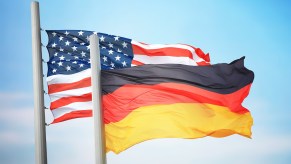 German flag against a blue sky, U.S. national flag in the background.