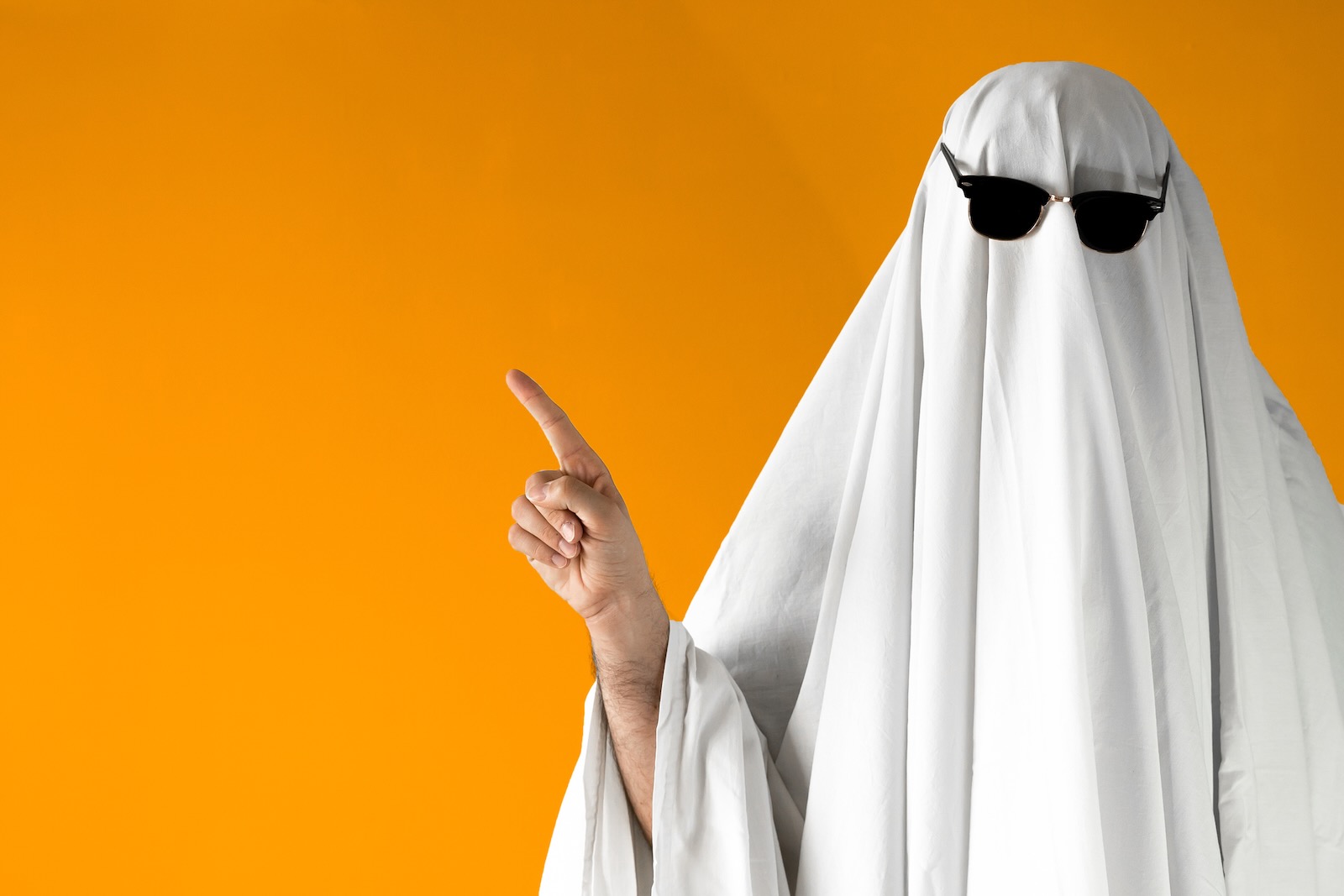 Person wearing a white sheet ghost costume and sunglasses stands in front of an orange background.