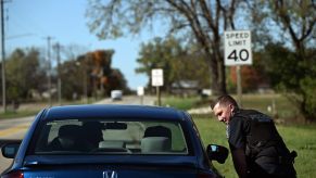 Police officer leans over to speak with a passenger in a blue Honda Civic during a traffic stop, trees visible in the background.