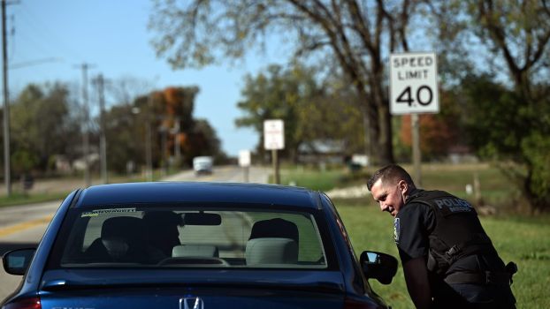 Police officer leans over to speak with a passenger in a blue Honda Civic during a traffic stop, trees visible in the background.