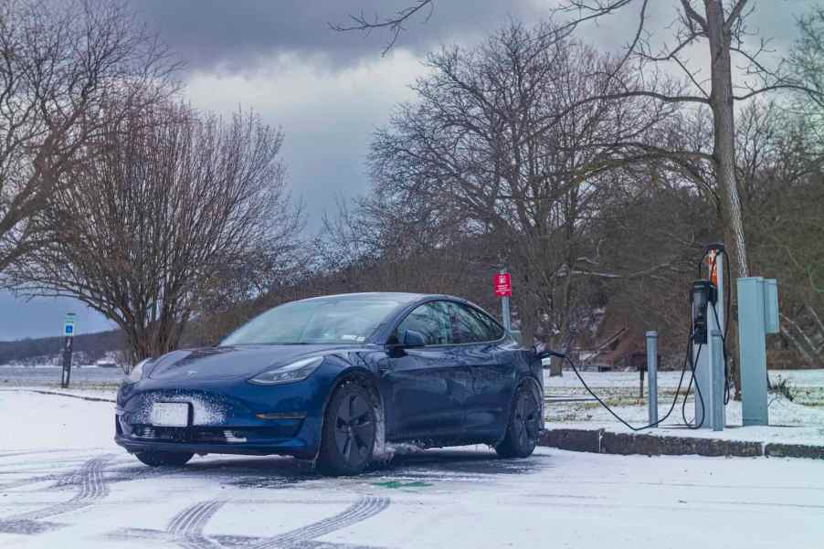 Blue Tesla Model 3 charging during the winter, snow visible in the foreground.
