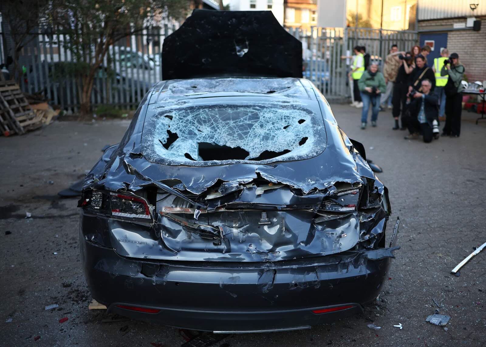 Blue Tesla sedan smashed during an Elon Musk protest, photographers in the background.