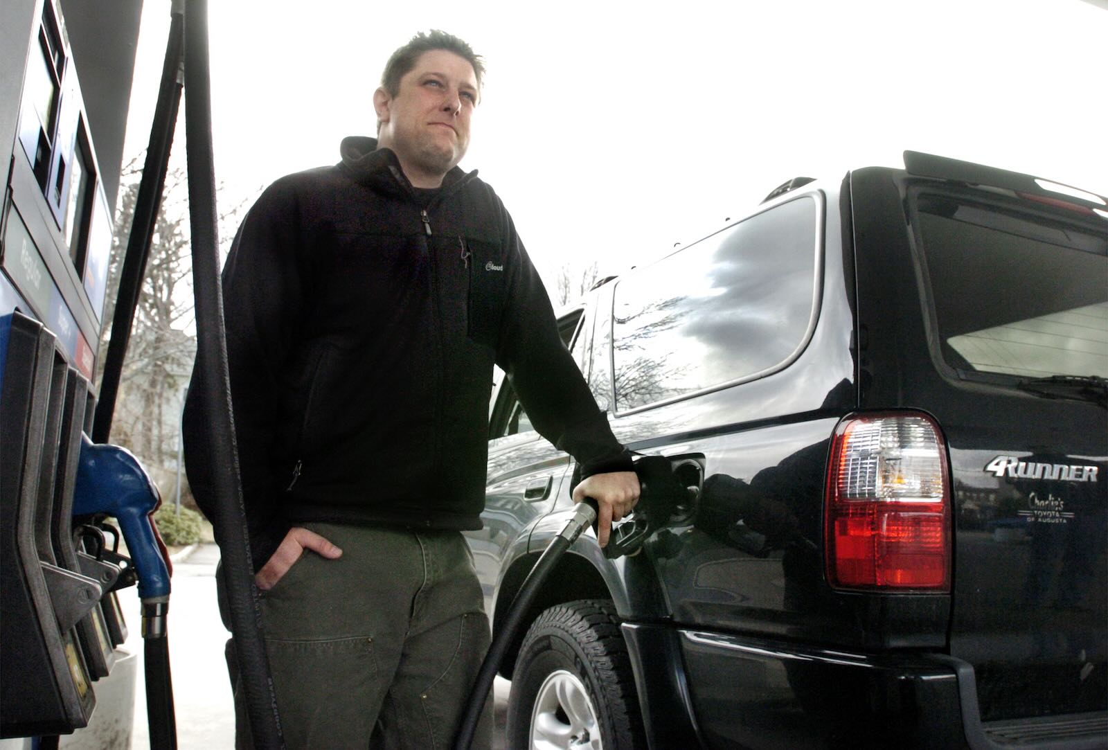 Man leans against his Toyota 4Runner while fueling it up at a gas station.