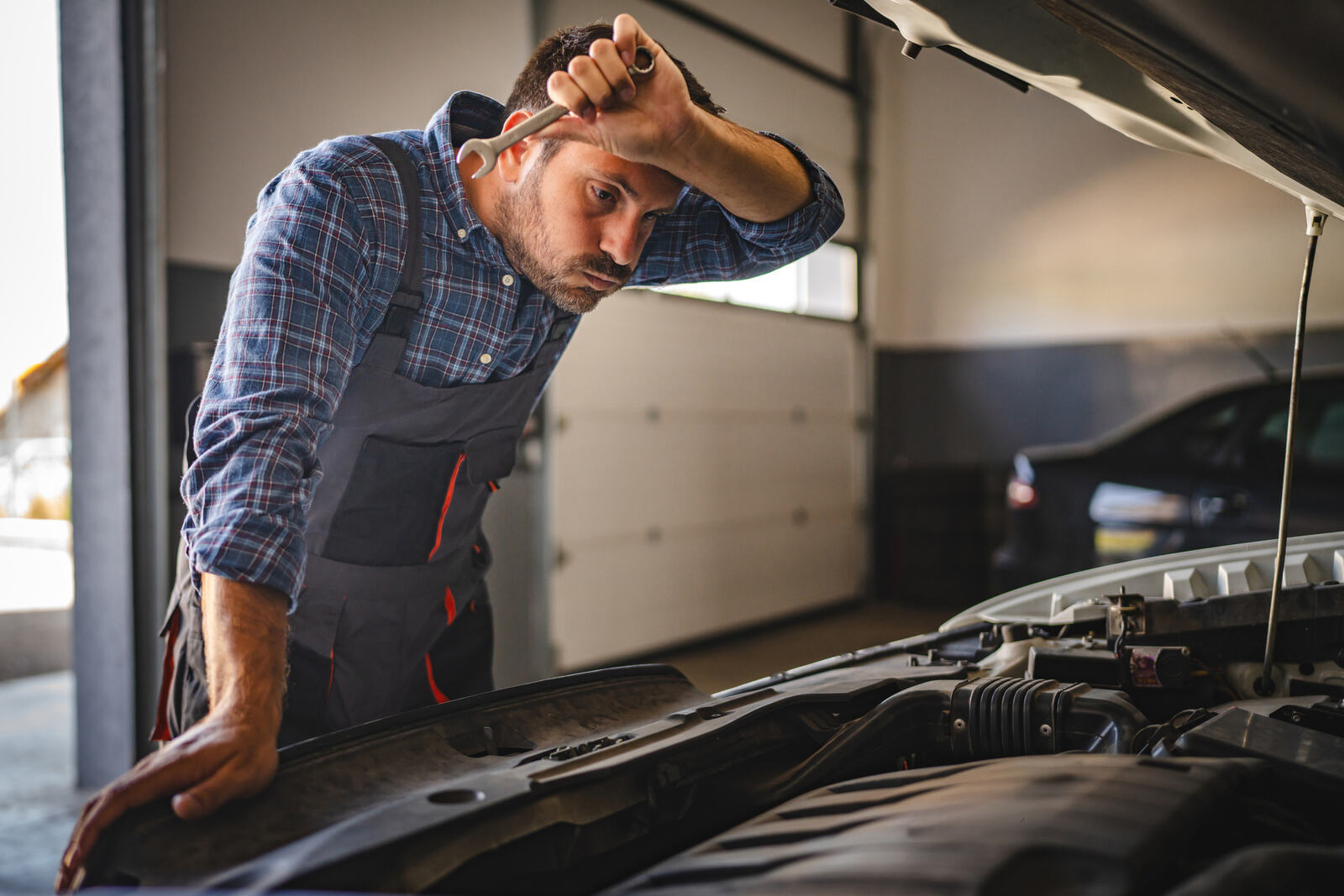 A frustrated mechanic leaning over an engine bay wipes forehead with arm