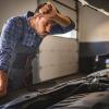 A frustrated mechanic leaning over an engine bay wipes forehead with arm