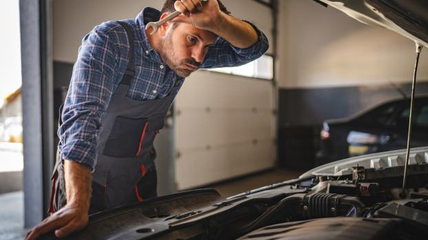 A frustrated mechanic leaning over an engine bay wipes forehead with arm