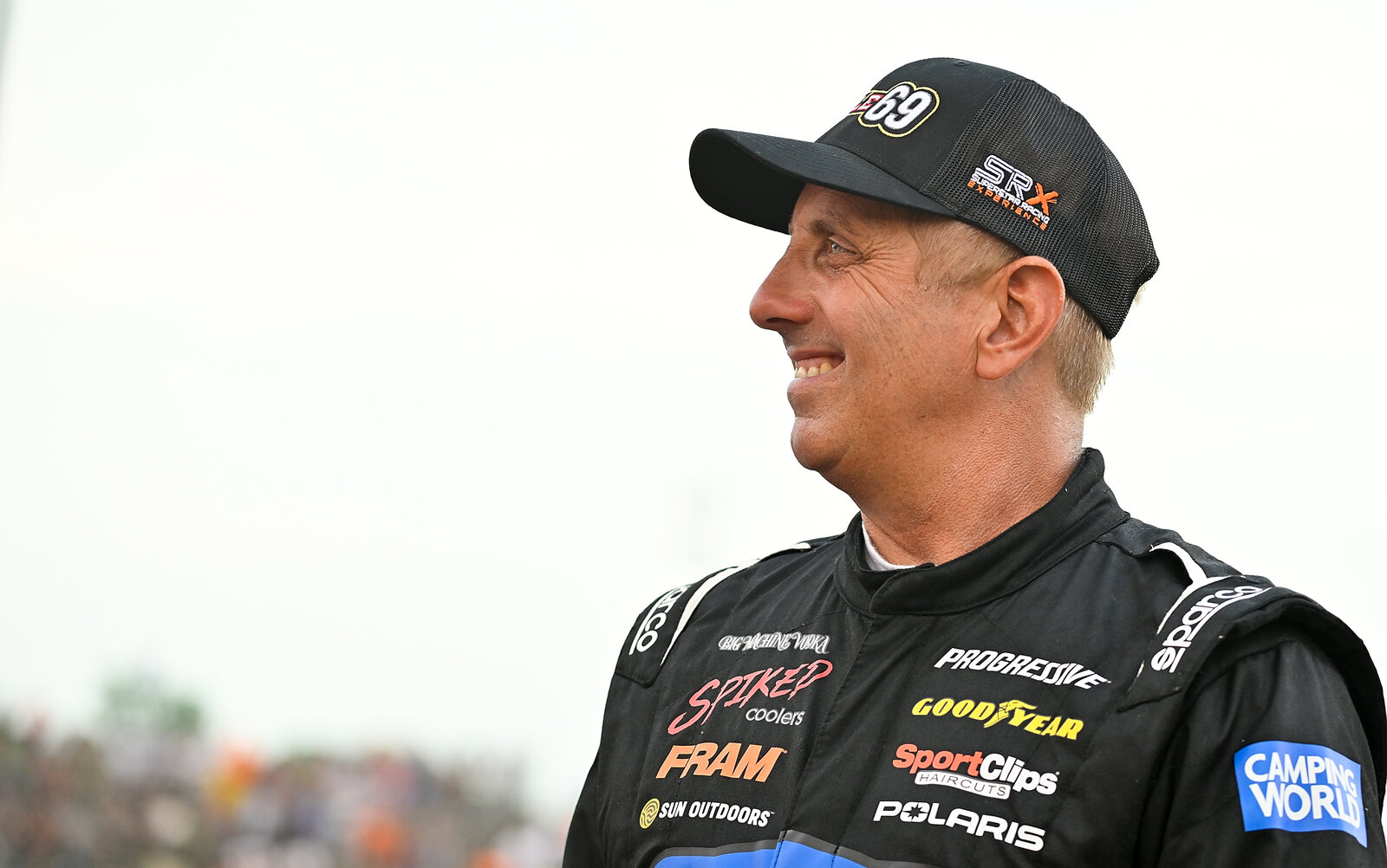 Greg Biffle, smiling, in race gear and baseball cap, looks to the left frame. View from chest up