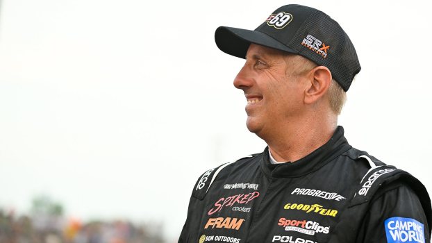 Greg Biffle, smiling, in race gear and baseball cap, looks to the left frame. View from chest up