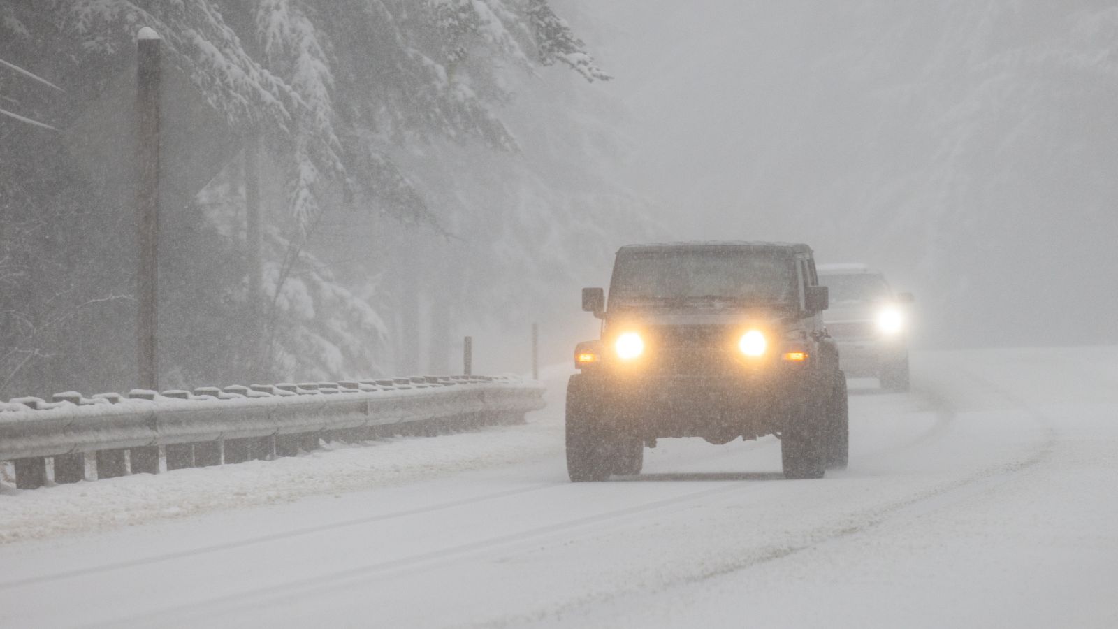 A Jeep with its headlights on drives on a snowy highway