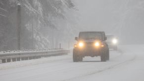 A Jeep with its headlights on drives on a snowy highway