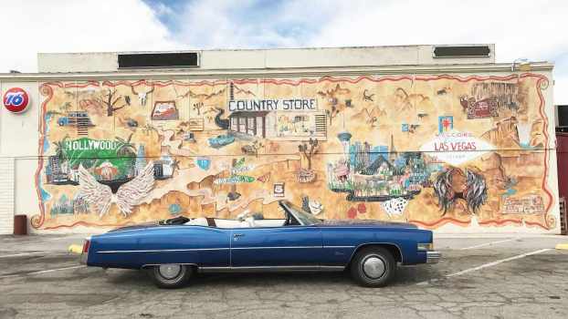 Blue Cadillac convertible parked in front of a mural on the side of a California store.