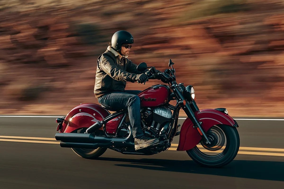 man in a leather jacket rides a retro-styled red Indian cruiser motorcycle down the highway.