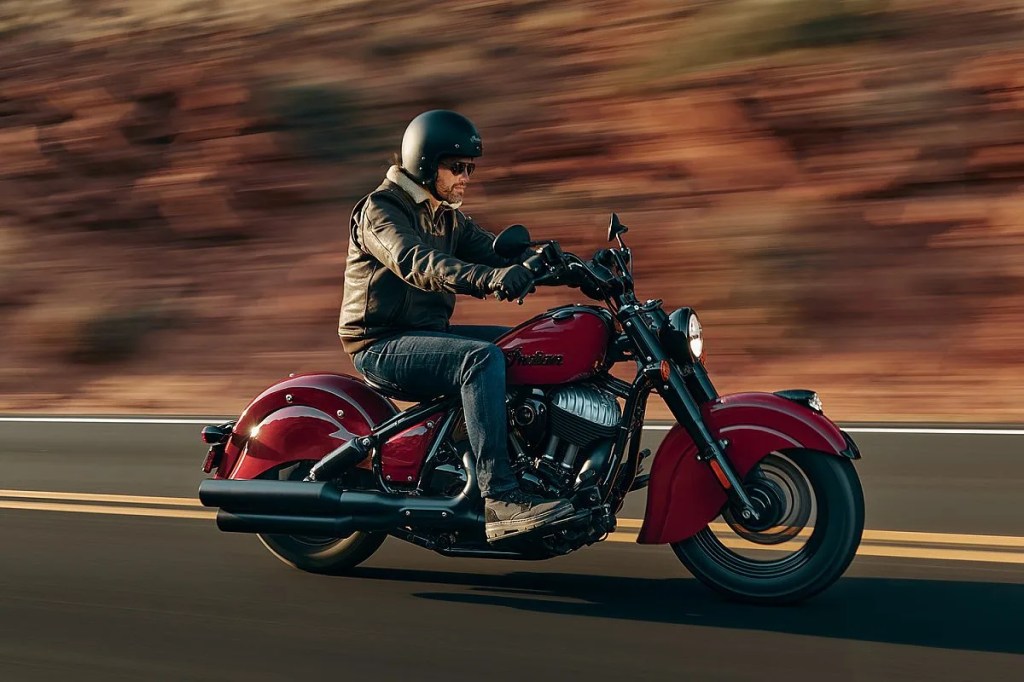 man in a leather jacket rides a retro-styled red Indian cruiser motorcycle down the highway.