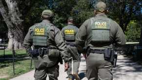 Three Federal agents in "Police: U.S. Border Patrol" vests walk through a park, trees in the background.
