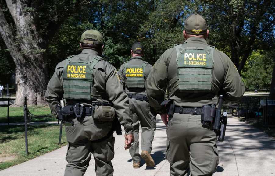 Three Federal agents in "Police: U.S. Border Patrol" vests walk through a park, trees in the background.