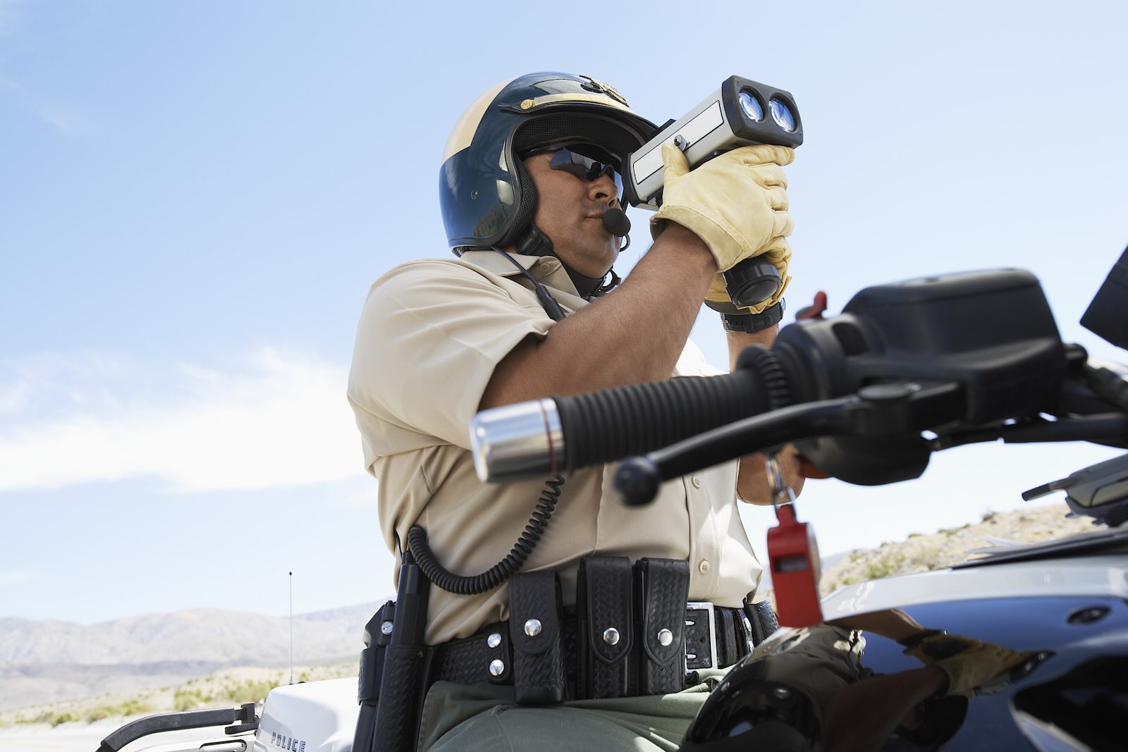 California Highway Patrol police officer standing over his motorcycle and holding a radar gun to detect speeders, blue sky visible behind him.