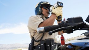 California Highway Patrol police officer standing over his motorcycle and holding a radar gun to detect speeders, blue sky visible behind him.