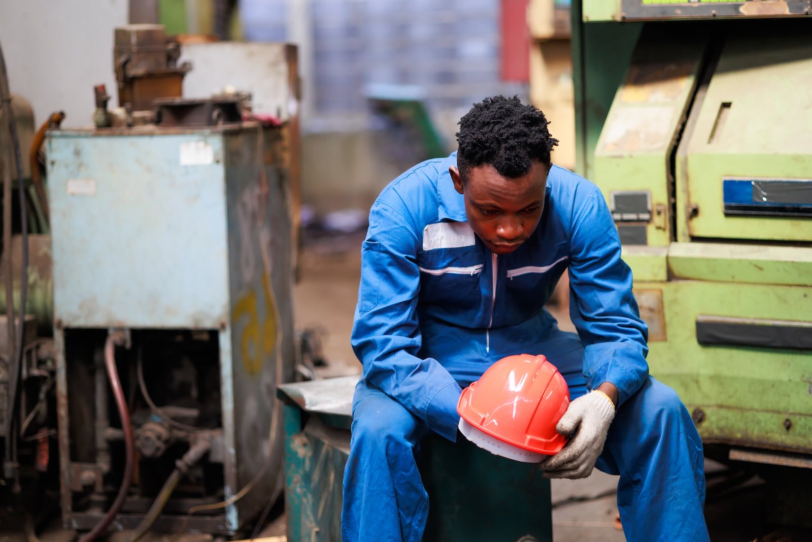 Worker sits in factory after layoffs, his orange helmet in his hands.