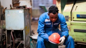 Worker sits in factory after layoffs, his orange helmet in his hands.