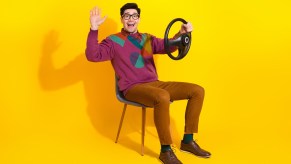 Friendly man smiles and waves, while holding a steering wheel in a regular chair