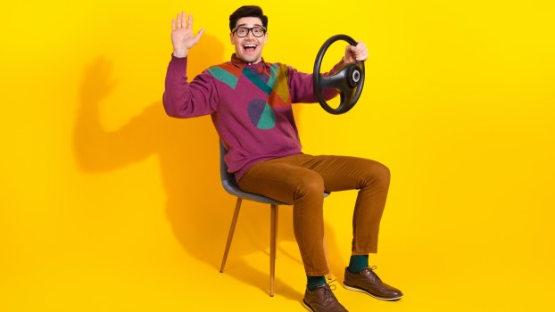 Friendly man smiles and waves, while holding a steering wheel in a regular chair