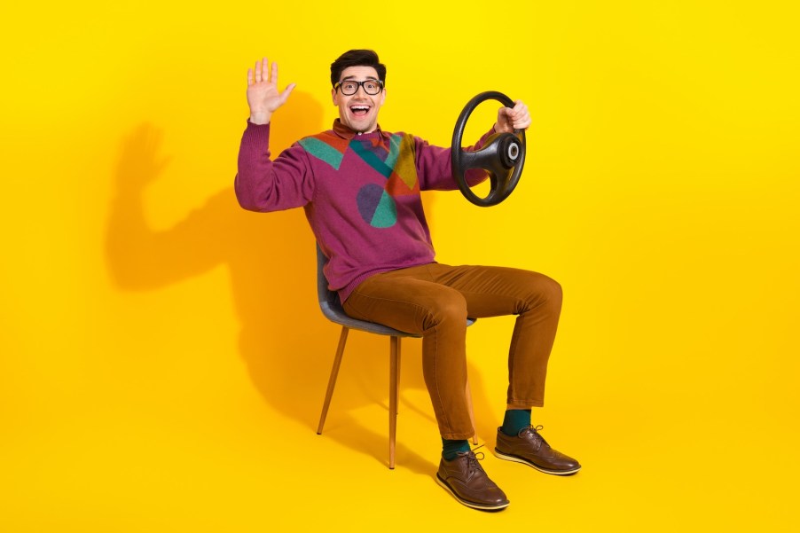 Friendly man smiles and waves, while holding a steering wheel in a regular chair