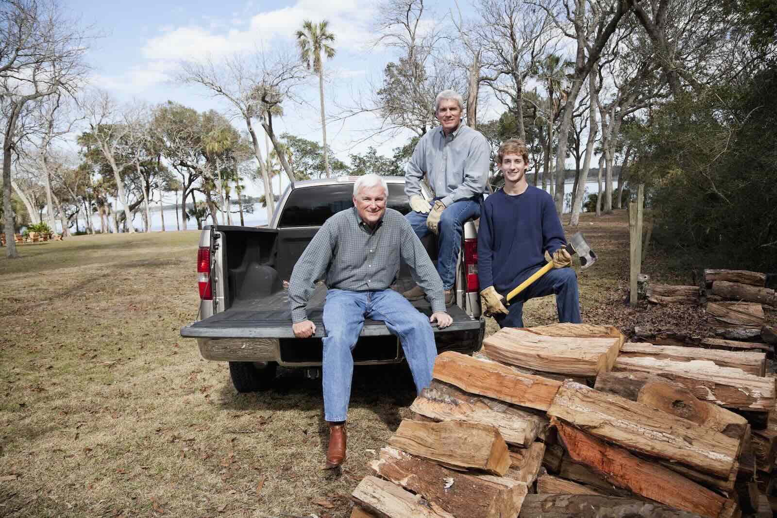 Three men sit in the bed of a pickup truck, a pile of split firewood in the foreground.