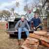 Three men sit in the bed of a pickup truck, a pile of split firewood in the foreground.