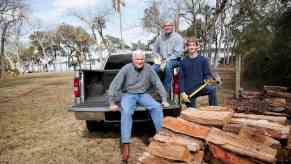 Three men sit in the bed of a pickup truck, a pile of split firewood in the foreground.