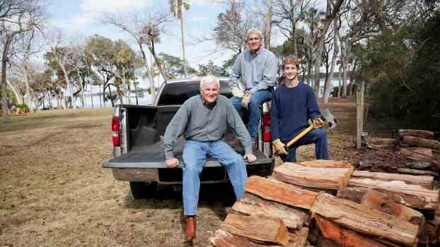 Three men sit in the bed of a pickup truck, a pile of split firewood in the foreground.