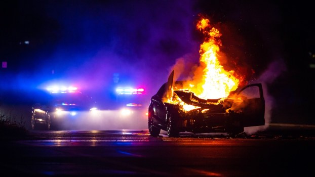 Yellow flames shoot from the engine compartment of a car stopped by police, blue lights in the background.