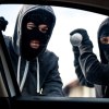 Two thieves stand outside a car, attempting to break its windows.