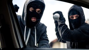 Two thieves stand outside a car, attempting to break its windows.