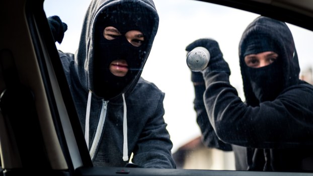 Two thieves stand outside a car, attempting to break its windows.