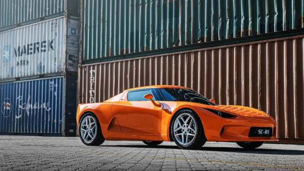 The front of an orange Chinese sports car in a harbor, shipping containers in the background.