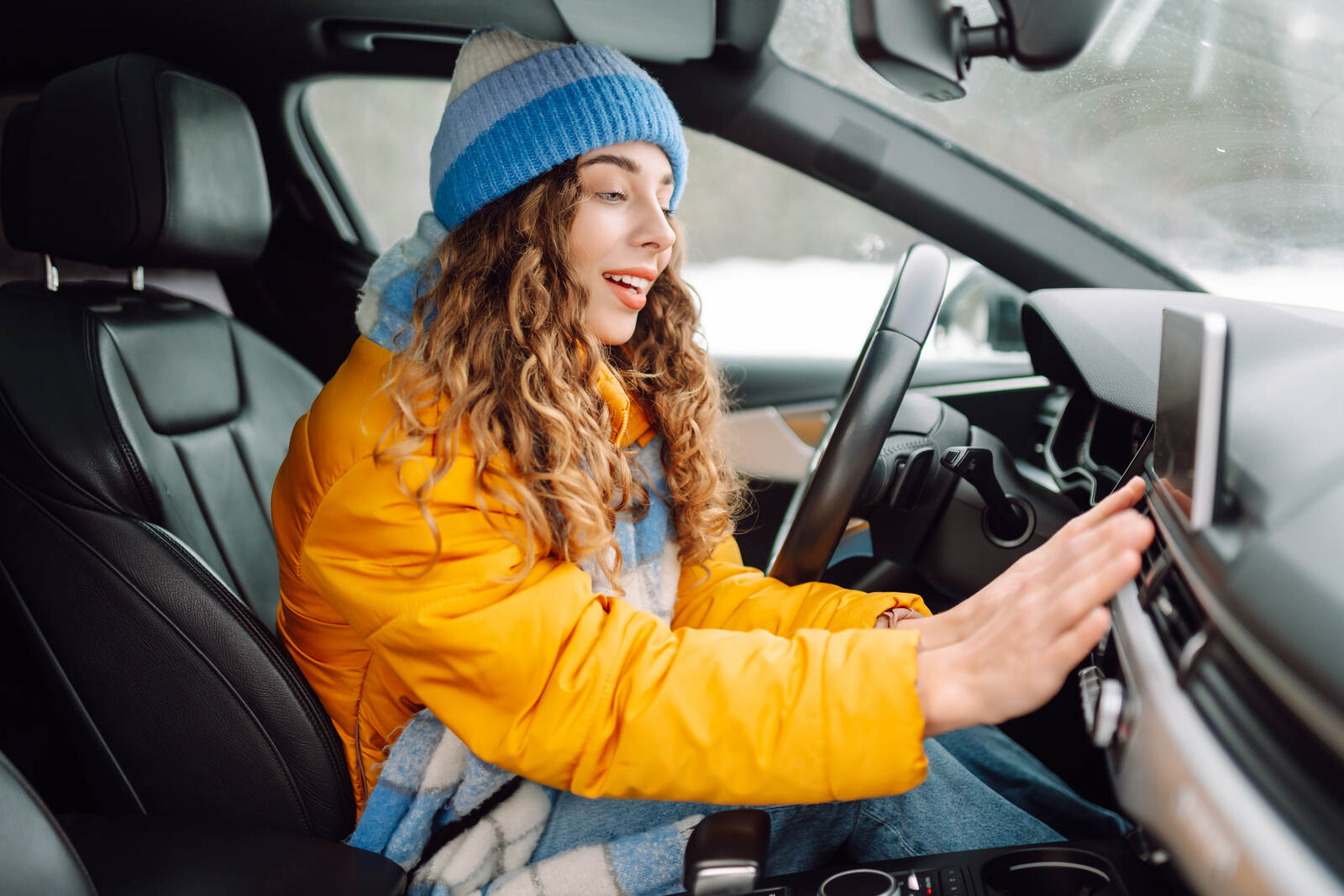 A female driver in winter coat and hat turns car heat on holds hand up to dash vent