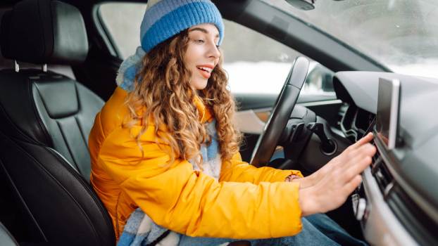 A female driver in winter coat and hat turns car heat on holds hand up to dash vent