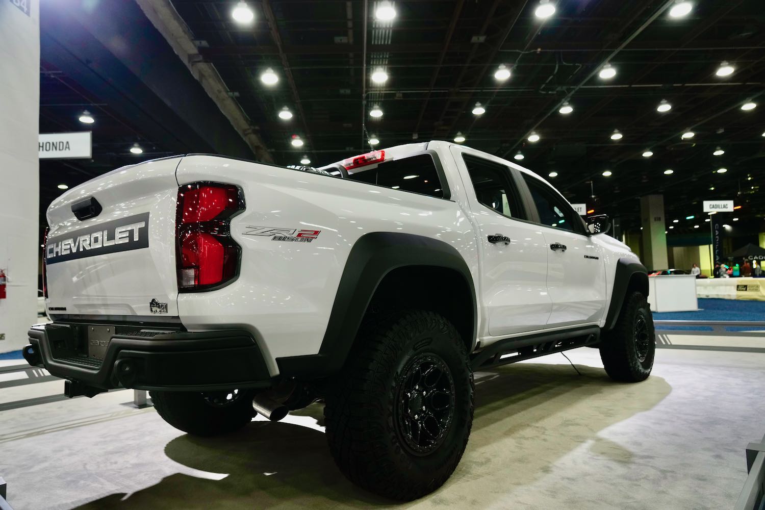 The bed and rear tire of a white Chevrolet Colorado midsize pickup truck parked at an auto show.