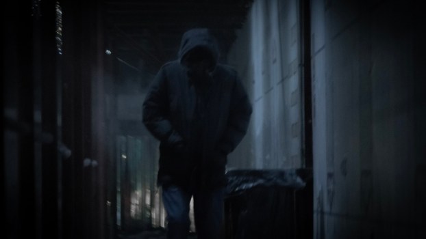 Man with a hood over his face walks through a dark alleyway