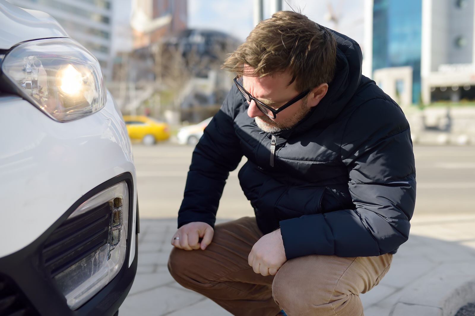 Man crouches by his white Toyota to check the turn signal is flashing, buildings visible in the background.