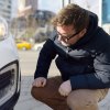 Man crouches by his white Toyota to check the turn signal is flashing, buildings visible in the background.