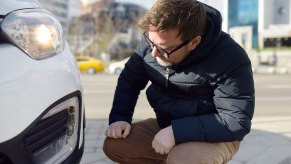 Man crouches by his white Toyota to check the turn signal is flashing, buildings visible in the background.