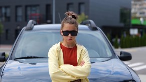 Gen Z woman leans against the hood of her car, a building visible in the background.